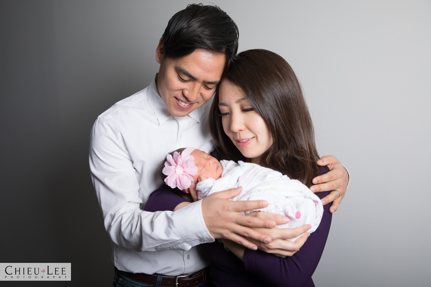 Half body parents mother mom father dad family embrace holding cuddling newborn baby girl sleeping eyes closed pink flower headband and white wrap white sweater light blue collar purple blouse gray gradient background