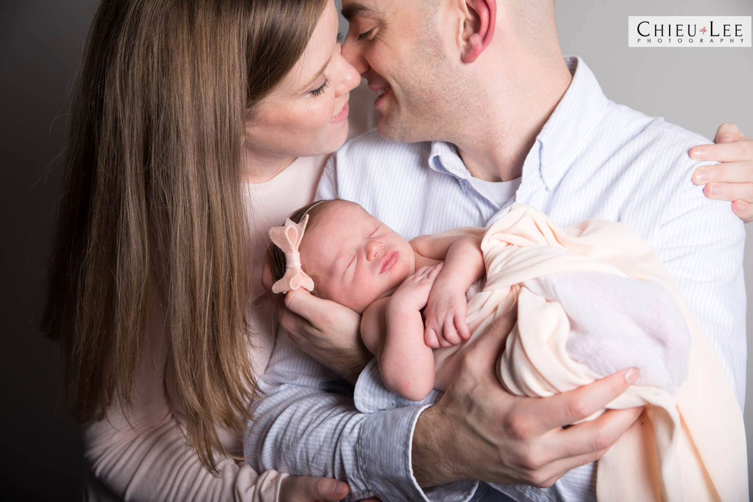 Closeup mother mom father dad kiss family cuddling holding embrace newborn baby girl sleeping eye closed pink bow headband ivory wrap light blue collared button-up shirt light pink sweater gray background