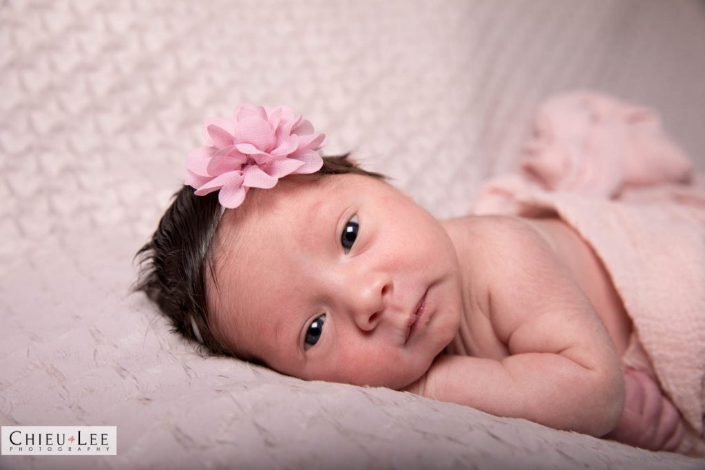 Closeup newborn baby girl awake eyes open pink flower headband and pink wrap on tan blanket