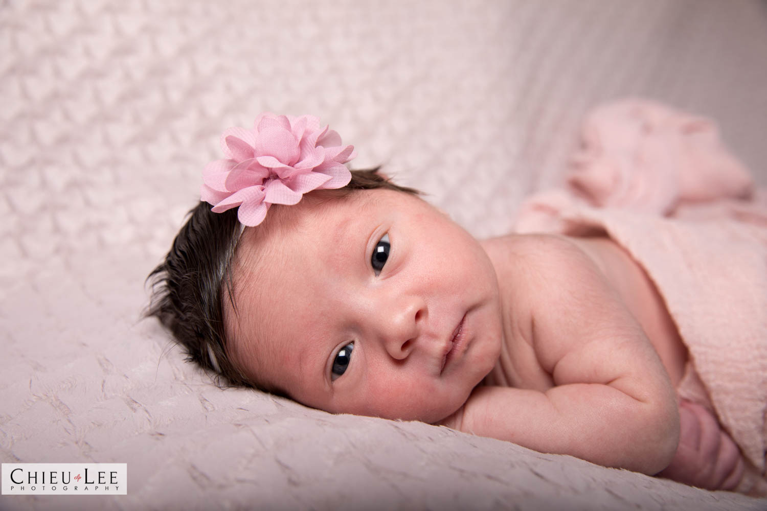 Closeup newborn baby girl awake eyes open pink flower headband and pink wrap on tan blanket