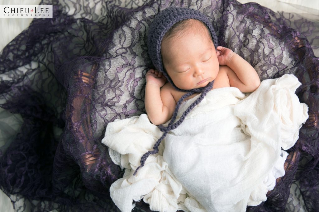 Newborn baby girl sleeping full body eyes closed purple crochet bonnet and white wrap on purple lace and white blanket