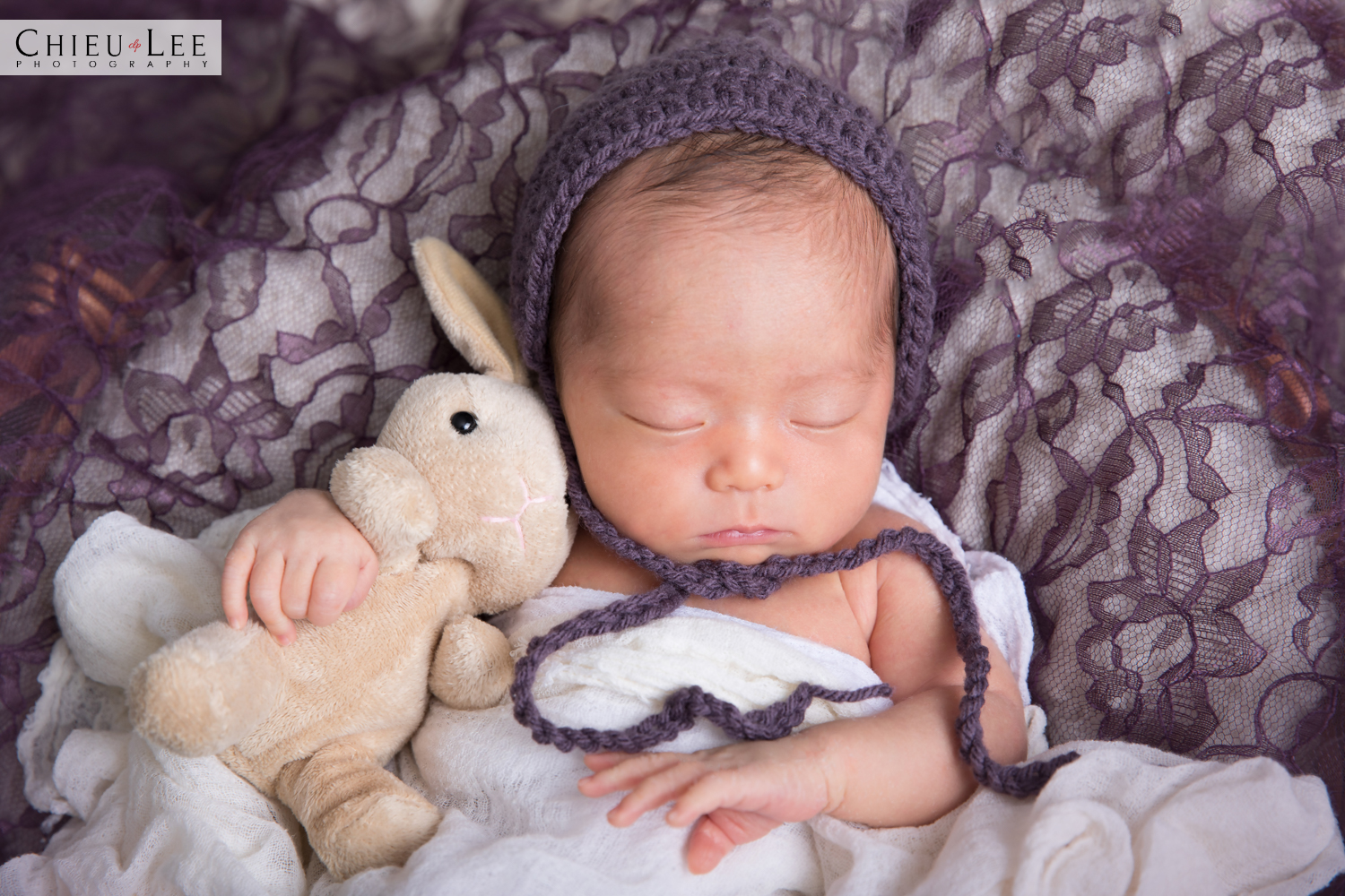 Newborn baby girl half body sleeping eyes closed purple crochet bonnet and white wrap cuddling beige plush bunny doll on purple lace and white blanket