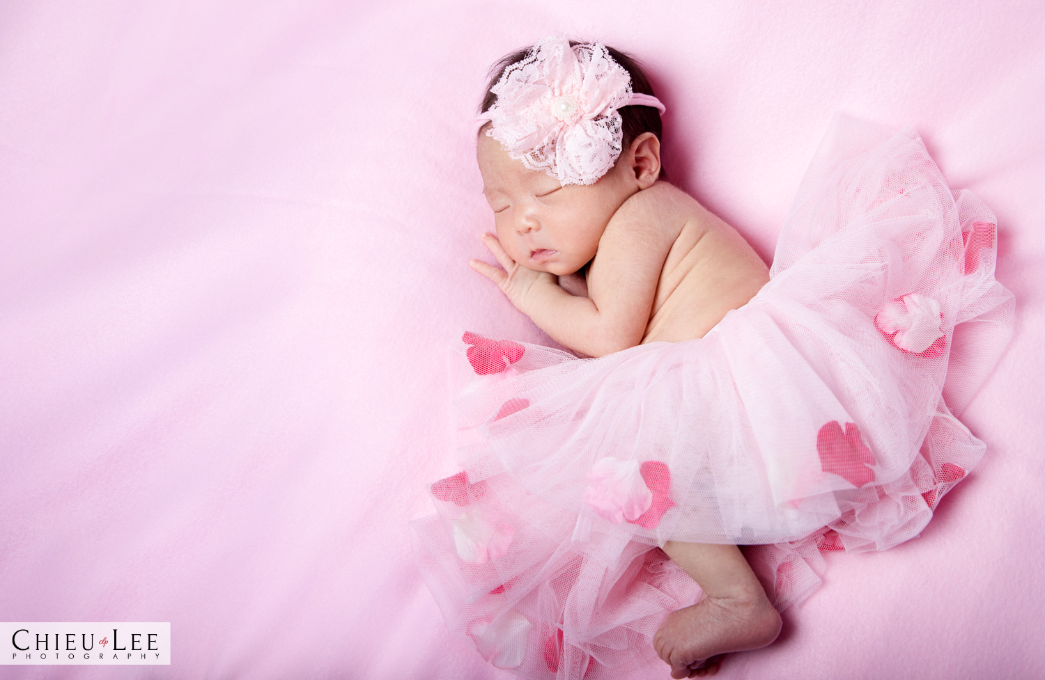 Newborn baby girl sleeping eyes closed full body lying on her side in a pink ballet tutu dress and white lace bow headband with flower petals